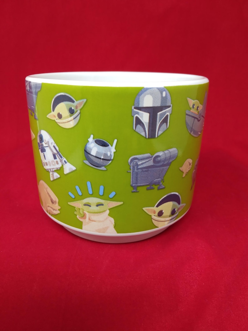 K&ouml;nnte beinhalten: Ein gr&uuml;ner Keramik-Blumentopf mit verschiedenen Star Wars-Charakteren, darunter The Mandalorian und Grogu. Der Topf hat eine wei&szlig;e Innenseite und einen roten Hintergrund.