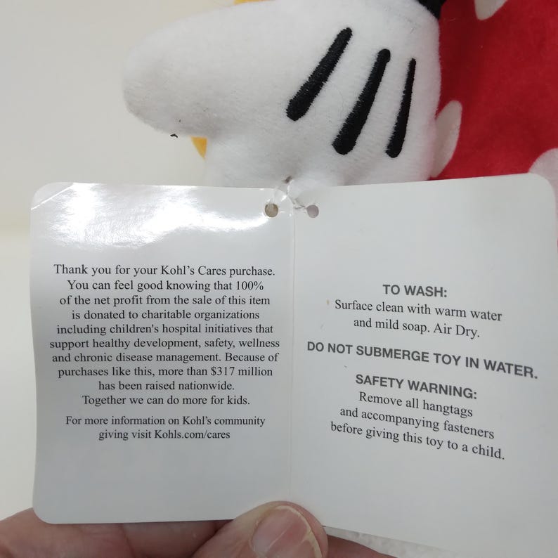 May include: A plush toy featuring a white glove with black details, part of a larger character. A tag is attached with text about charitable donations and care instructions. The toy is red, white, and black.