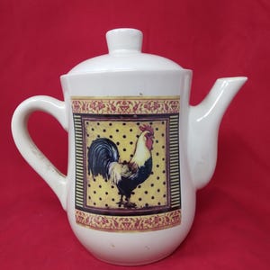 May include: White ceramic teapot with a lid and a curved handle. The teapot features a decorative panel with a rooster illustration on a yellow background with black polka dots. The panel is framed with a floral design.