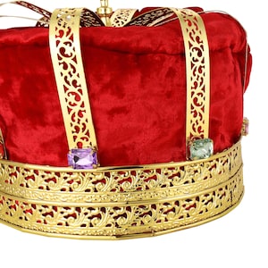 May include: Close-up of a king's crown. The crown is red velvet with gold metal filigree detailing.  The gold accents feature ornate cut-outs.  Purple and green jewel-like accents are set into the gold metal band. The crown is a festive accessory perfect for royalty, theater, or costume parties.