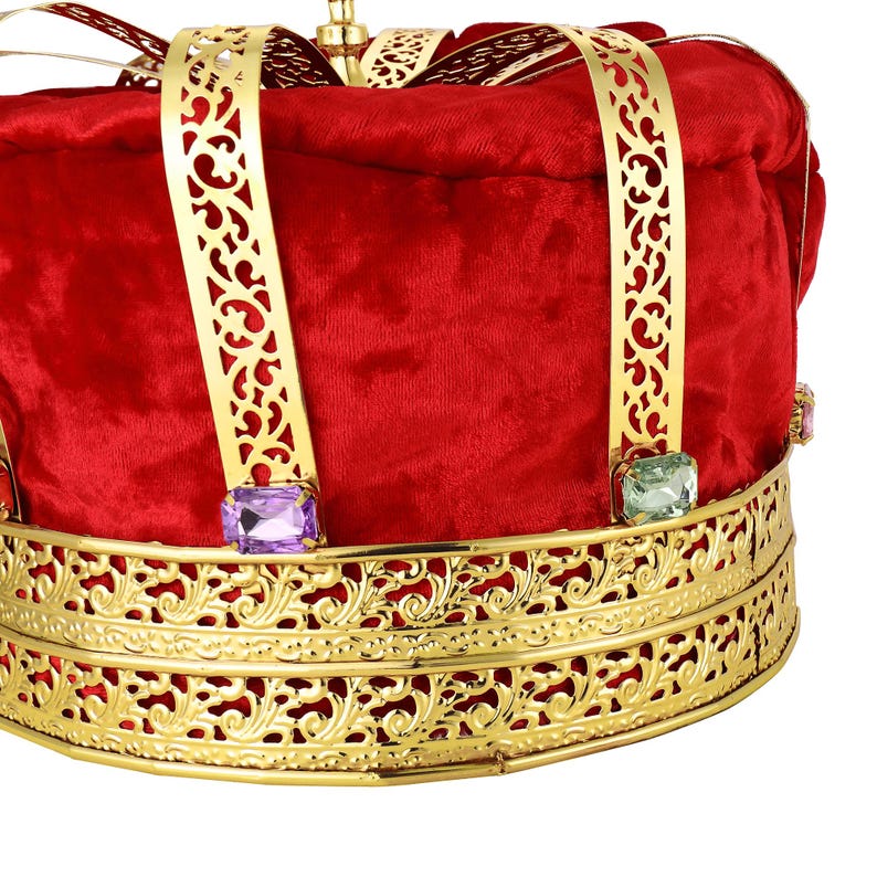 May include: Close-up of a king's crown. The crown is red velvet with gold metal filigree detailing.  The gold accents feature ornate cut-outs.  Purple and green jewel-like accents are set into the gold metal band. The crown is a festive accessory perfect for royalty, theater, or costume parties.
