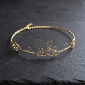 May include: A delicate gold-colored wire crown or headband with an intricate, looping design. The jewelry piece is displayed on a dark, textured surface, showcasing its elegant and ornamental style. The crown's design features small loops and swirls.