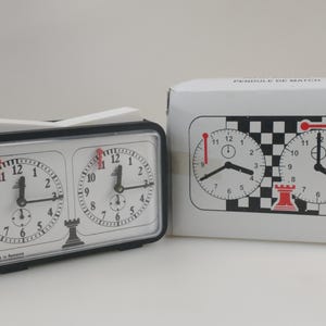 May include: A black and white chess clock with two clock faces, red hands, and a white switch on top. The clock is next to a white box with a chess clock graphic. The clock face has the text "Made in Romania".