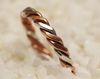 May include: A twisted silver and copper ring, with a smooth, polished finish. The ring is photographed on a bed of white stones.