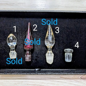 May include: Four glass bottle stoppers in a black box. The stoppers are numbered 1 through 4. Stopper 1 is clear glass with a round top. Stopper 2 is a deep purple glass with a pointed top. Stopper 3 is clear glass with a pointed top. Stopper 4 is a clear glass cylinder. Stoppers 1, 2, and 3 are marked "Sold".