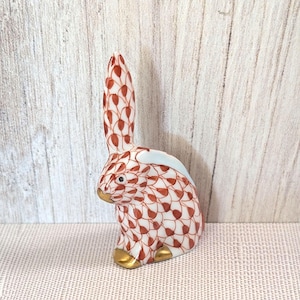 May include: A ceramic rabbit figurine with a white base and a red leaf pattern. The rabbit has gold-colored feet and a gold-colored nose. The ears are long and upright. The figurine is on a light pink surface.
