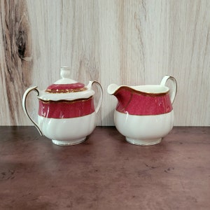 May include: A white porcelain sugar bowl and creamer set with a red and gold floral design. The sugar bowl has a lid.