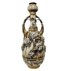 Op de afbeelding: Een hoge, decoratieve keramische vaas met een crèmekleurige basis en een donkerbruine bovenkant. De vaas heeft twee handvatten en is versierd met witte bloemversieringen en gouden accenten. De vaas is ongeveer 61 cm hoog.