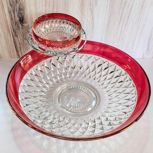 May include: A clear glass serving bowl with a red rim and a smaller matching bowl on a metal stand. Both bowls have a diamond pattern.