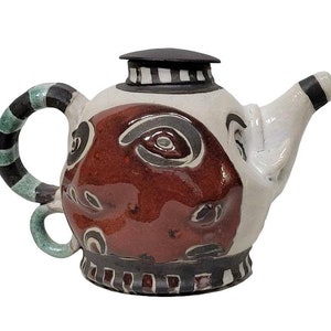 Può includere: Una teiera in ceramica bianca con un coperchio a strisce bianche e nere e un manico a strisce verdi e nere. La teiera è decorata con un viso rosso, nero e bianco.
