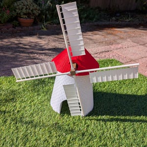 May include: A white windmill model with a textured brick pattern on the base and a bright red conical roof. The structure features four white rectangular blades attached to a central brown hub, creating a detailed miniature representation of a traditional windmill.
