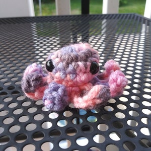 May include: A small crocheted octopus with pink, purple, and gray yarn. The octopus has black eyes and is sitting on a black metal surface.