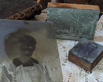 May include: A collection of antique printing blocks and a vintage photograph are displayed on a lace cloth. The blocks depict architectural scenes and a portrait. A leather-bound book is in the background, hinting at a historical or artistic context.
