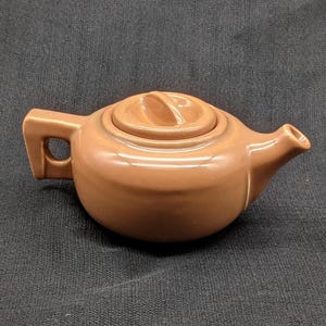 May include: A small, brown ceramic teapot with a matching lid. The teapot has a rounded body, a handle with a rectangular opening, and a spout. The lid has a small handle for easy removal. The teapot is likely used for brewing tea.