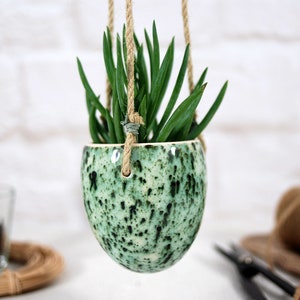 Puede incluir: Un macetero colgante de cerámica verde moteado con una planta suculenta en su interior. El macetero está suspendido de una cuerda de yute natural.