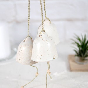 May include: A set of three white ceramic bells with a speckled finish, hanging from twine with wooden beads. The bells are suspended against a white brick background.