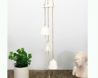 May include: A white ceramic wind chime with three bells hanging from a string. The bells are decorated with small holes and the string is made of natural fiber.