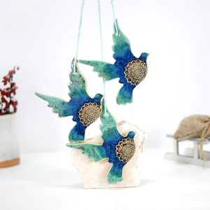 May include: Three ceramic doves with blue and green glaze and gold accents hanging from a white string. The doves are arranged in a cluster and are suspended from a white surface.