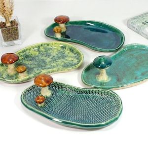May include: A selection of ceramic serving dishes in shades of green and blue, each featuring a mushroom-shaped handle. The dishes have varied textures and shapes, with a glossy finish. A book with Polish text is visible in the background.