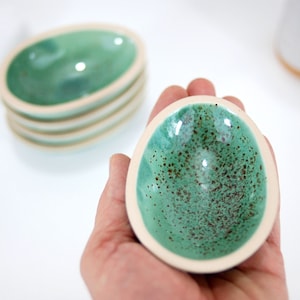 Può includere: Un piccolo piatto in ceramica di forma ovale con una glassa verde macchiata e un bordo bianco. Il piatto è tenuto in una mano.