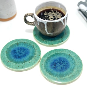 Puede incluir: Tres posavasos redondos de cerámica con un diseño de remolino azul y verde. Los posavasos están sobre una superficie blanca.