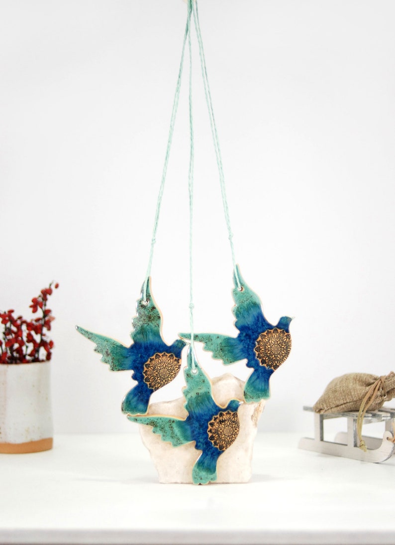 May include: Three ceramic blue and green birds with gold accents hanging from a string. The birds are in flight and have a mandala design on their chests.