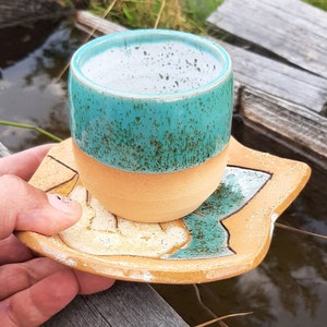 Op de afbeelding: Een teal en beige keramische mok met een gespikkeld glazuur staat op een bijpassende schotel. De schotel heeft een bladontwerp.