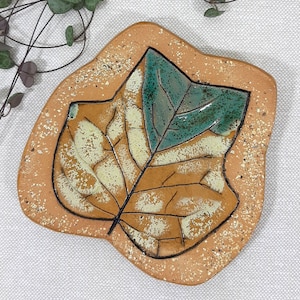 Op de afbeelding: Een keramische schaal in de vorm van een blad met een bruine en groene glazuur. Het blad heeft een gedetailleerd adernetwerk en een gespikkelde textuur.