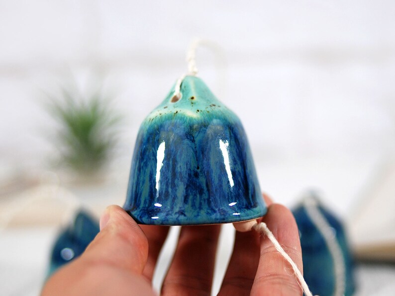 May include: A blue and green ceramic wind chime with a bell-shaped design. The chime is hanging from a white string.
