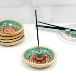 Peut inclure: Un porte-encens en c&eacute;ramique bleu-vert avec un motif de mandala rouge et or. Le porte-encens a un petit c&ocirc;ne en c&eacute;ramique bleu-vert au centre pour br&ucirc;ler des b&acirc;tonnets d'encens. Trois c&ocirc;nes d'encens en c&eacute;ramique bleu-vert sont sur une assiette en verre transparent &agrave; l'arri&egrave;re-plan.