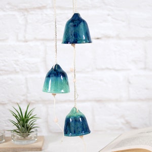 May include: A set of three ceramic bells hanging from a string. The bells are blue and green with a gradient effect. The bells are hanging in front of a white brick wall.