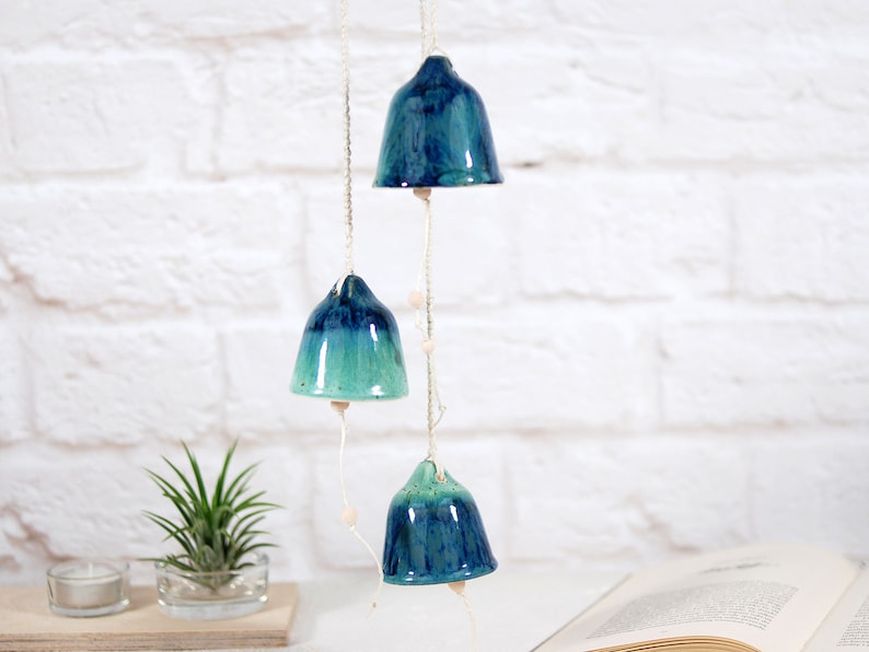 May include: A set of three ceramic bells hanging from a string. The bells are blue and green with a gradient effect. The bells are hanging in front of a white brick wall.