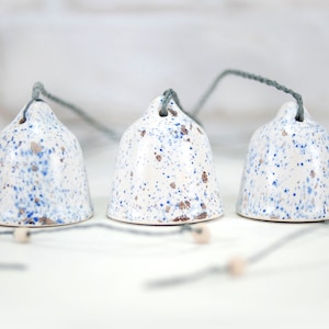 May include: Three white ceramic bells with blue and gold speckles hang from gray strings. The bells are tied together in a cluster.