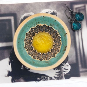 Peut inclure: Un petit plat en c&eacute;ramique avec un gla&ccedil;age bleu sarcelle et un motif de mandala noir et blanc. Le centre du mandala est un cercle jaune.