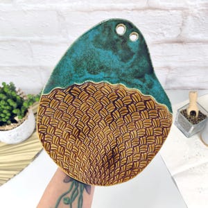 Könnte beinhalten: Handgefertigte Keramik-Servierplatte mit einzigartigem Design. Der obere Teil ist tief türkis, während der untere Teil ein geflochtenes Korbmuster in Braun- und Goldtönen aufweist. Die Platte hat zwei kleine Löcher oben und ist etwa 25 cm lang.
