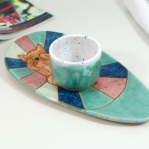 May include: A teal ceramic serving dish with a geometric design featuring a cat illustration. A small teal and white ceramic bowl sits on the dish.