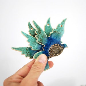 May include: A ceramic wall hanging in the shape of a blue and green bird with a gold mandala design. The bird is in flight with its wings spread out.
