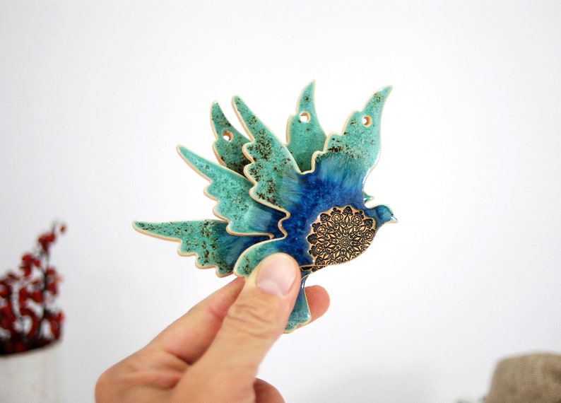 May include: A ceramic wall hanging in the shape of a blue and green bird with a gold mandala design. The bird is in flight with its wings spread out.