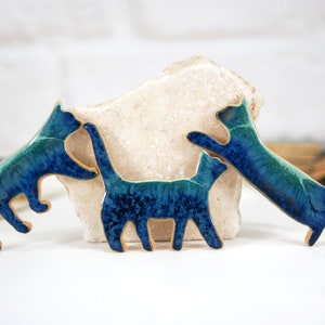 Può includere: Tre figurine di gatto in ceramica in tonalità blu e verdi. I gatti sono in diverse pose, uno in piedi sulle zampe posteriori, uno seduto e uno sdraiato.