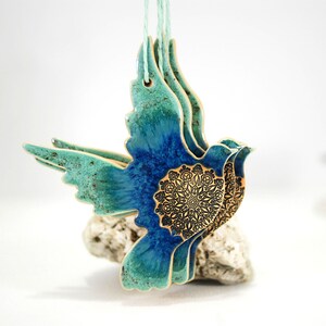 May include: A ceramic dove ornament with a blue and green glaze and a gold mandala design. The dove is in flight and has a rustic, handmade look.
