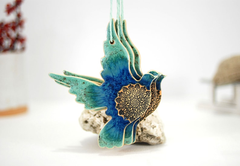 May include: A ceramic dove ornament with a blue and green glaze and a gold mandala design. The dove is in flight and has a rustic, handmade look.