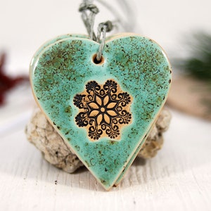 May include: Teal ceramic heart ornament with a black snowflake design. The ornament has a rustic, distressed look and is strung with a twine cord.
