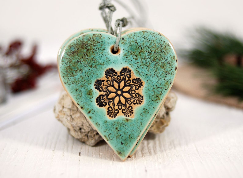 May include: Teal ceramic heart ornament with a black snowflake design. The ornament has a rustic, distressed look and is strung with a twine cord.