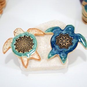May include: Two ceramic turtle figurines with mandala designs. The turtle on the left is light brown with a teal mandala, and the turtle on the right is blue with a gold mandala.