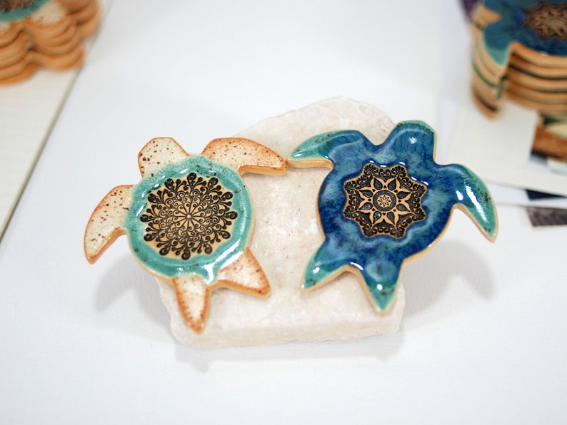 May include: Two ceramic turtle figurines with mandala designs. The turtle on the left is light brown with a teal mandala, and the turtle on the right is blue with a gold mandala.