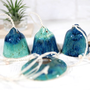 May include: Four blue and green ceramic bells with white string hanging from them. The bells are hanging on a white surface.