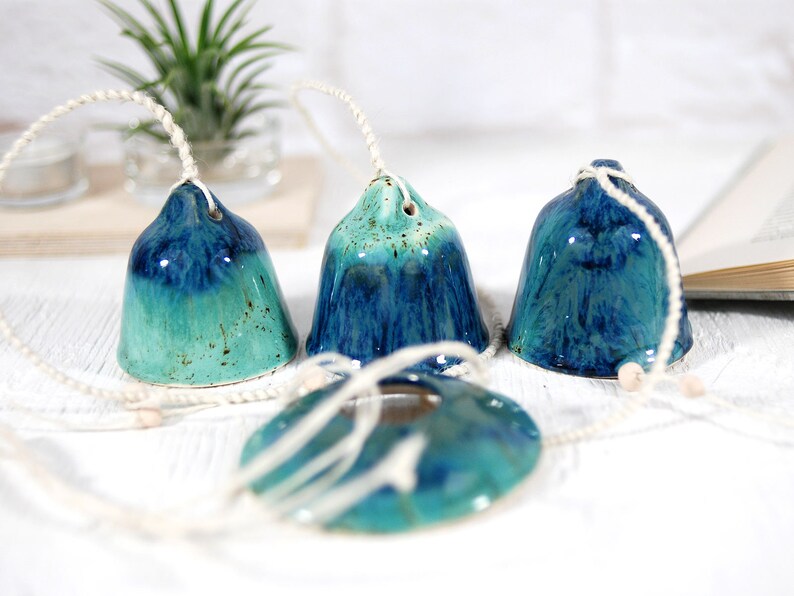 May include: Four blue and green ceramic bells with white string hanging from them. The bells are hanging on a white surface.