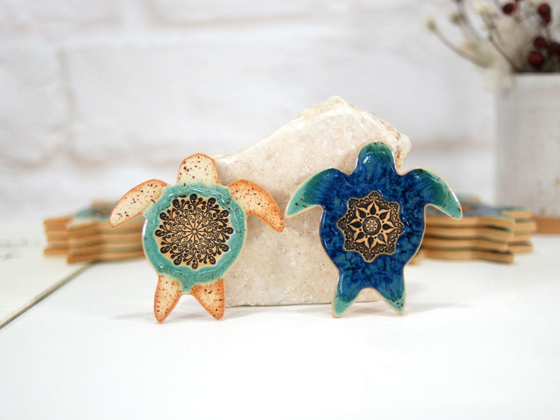 May include: Two ceramic turtle figurines with mandala designs. The turtle on the left is light brown with a teal and black mandala. The turtle on the right is blue with a gold and black mandala.