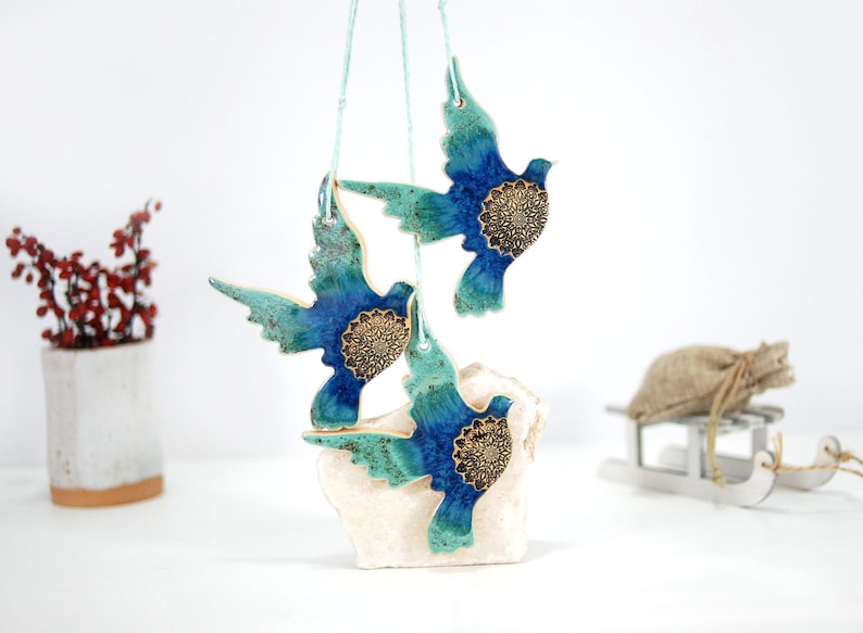 May include: Three ceramic doves with blue and green glaze and gold accents hanging from a white string. The doves are arranged in a cluster and are suspended from a white surface.