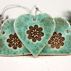 May include: Three teal blue ceramic heart ornaments with black snowflake designs. Each heart has a string for hanging.